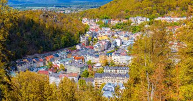 Karlovy Vary şehrinin rengarenk binaları ve tarihi merkezlerinde spa otelleri olan panoramik manzarası. Karlsbad kasabası ve Slavkov Ormanı tepeleri Sonbaharda yamaçta ağaçlarla