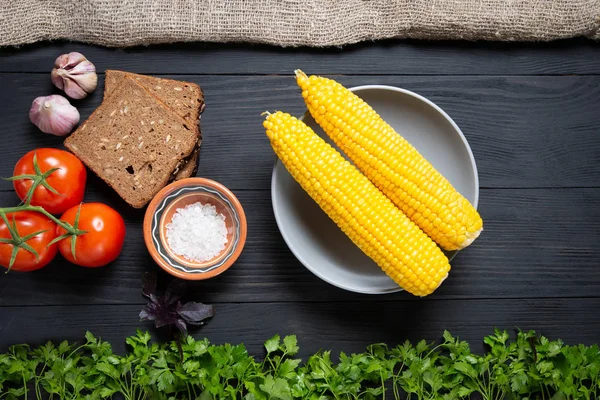 plate of corn on a black wooden background with lots of fresh vegetables around