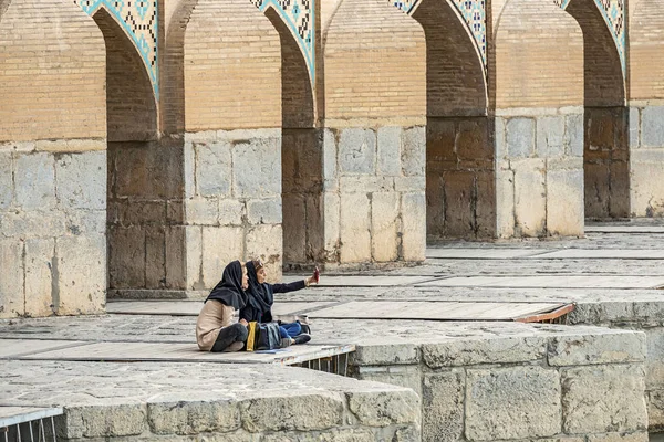 22/05/2019 Isfahan, Iran, two iranian girls is sitting on Khaju Bridge with plenty of arches over Zayandeh river, and make a selfie on smartphone