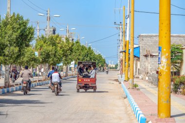 05/11/2019 Hürmüz, Hürmozgan Eyaleti, İran, elleri ile el sallayan neşeli insanlarla dolu caddeden aşağı yaslı bir motosiklet