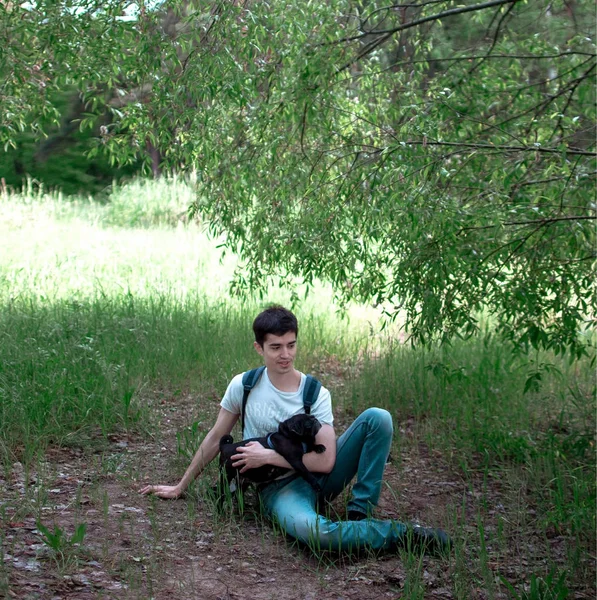 A young guy is resting under a tree with his black pug