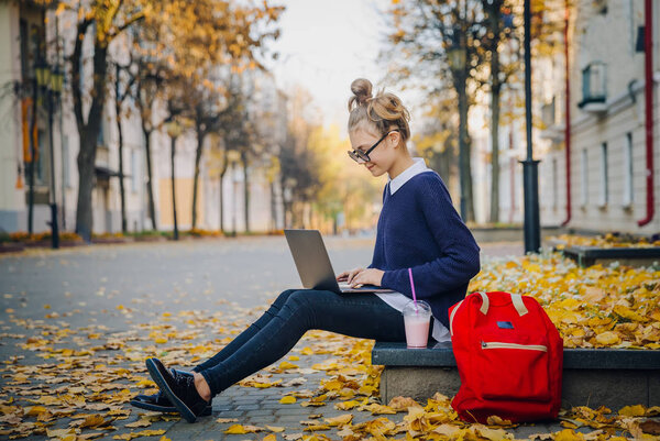 Pretty hipster teen girl sitting on a sidewalk on autumn city street and working laptop computer. Schoolgirl using notebook outdoor. Beautiful autumn weather.