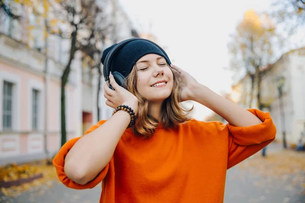 Jovem menina adolescente bonita hipster em chapéu preto ouvindo música via fones de ouvido na rua de outono — Fotografia de Stock