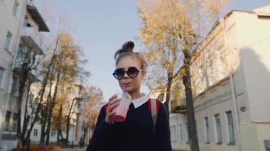 Güzel hipster teen kırmızı çanta ile bina arasında street yürüyüş plastik bir kap dan muzlu süt içiyor. Güneş gözlüklü şirin kız bir saman içecek içecekler.