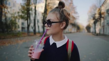 Güzel hipster teen kırmızı çanta ile bina arasında street yürüyüş plastik bir kap dan muzlu süt içiyor. Güneş gözlüklü şirin kız bir saman içecek içecekler.