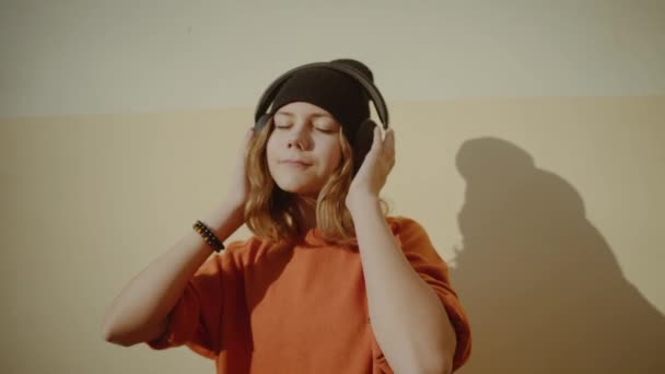 Carino giovane ragazza ascoltando musica in cuffia, stile urbano, elegante hipster teen in cappello nero ascoltare musica e sorridere davanti al muro giallo, arancione stile strada pazzo — Video Stock