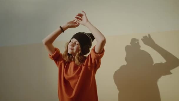 Schattig jong meisje luisteren muziek in hoofdtelefoons en dansing, stedelijke stijl, stijlvolle hipster tiener in zwarte hoed luisteren muziek en glimlach infront van gele muur, oranje gekke Streetstyle — Stockvideo