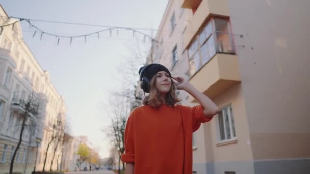 Söt ung flicka gick nedför gamla staden gatan och lyssna musik i hörlurar, urban stil, snygg hipster tonåring i svart hatt lyssna musik och leende, orange galna stil — Stockvideo