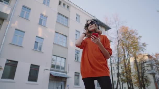 Söt ung flicka lyssnar musik i hörlurar, urban stil, snygg hipster tonåring i svart hatt lyssna musik och leende infront av oidsity byggnader, orange galna gatan stil — Stockvideo