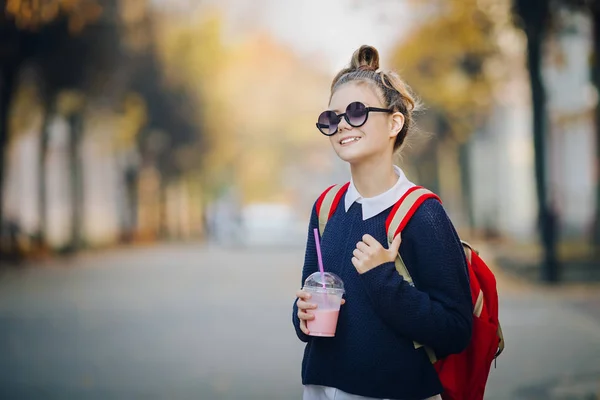 Güzel hipster teen kırmızı çanta ile bina arasında street yürüyüş plastik bir kap dan muzlu süt içiyor. Güneş gözlüklü şirin kız bir saman içecek içecekler.