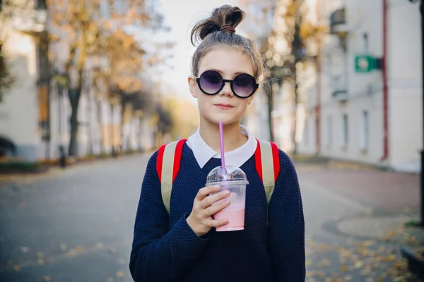 赤いバッグとかなり流行に敏感なティーンは、プラスチック カップの建物の間の通りを歩いてからミルクセーキを飲みます。サングラスでかわいい女の子がストローで飲み物を飲む. — ストック写真