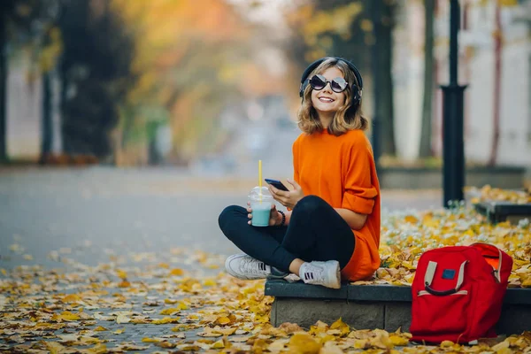 Jovem menina adolescente bonita hipster ouvir música via fones de ouvido, sentado em uma calçada na rua da cidade de outono e dinking palha de tromuto batido — Fotografia de Stock