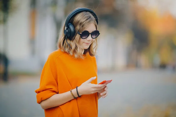 Aranyos fiatal lány sétál a régi város utca és a hallgató zene fejhallgató, városi stílusú, elegáns csípő tini hold mozgatható telefon és és őrült stílust mosoly, narancs — Stock Fotó