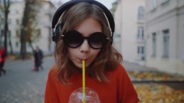 Güzel hipster teen dinlemek müzik içecekler milkshake binalar arasında street yürüyüş plastik bir kap dan kulaklık, kapatın. Güneş gözlüklü şirin kız bir saman içecek içecekler.