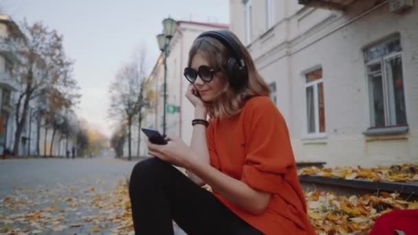 Roztomilá mladá dívka poslech hudby sluchátka, městský styl, stylový bederní teen sedí na chodníku na ulici a volba skladby na mobilní telefon infront z oidsity budov, oranžové ulice — Stock video