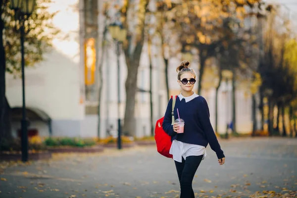 Güzel hipster teen kırmızı çanta ile bina arasında street yürüyüş plastik bir kap dan muzlu süt içiyor. Güneş gözlüklü şirin kız içecekler içmek bir saman. — Stok fotoğraf