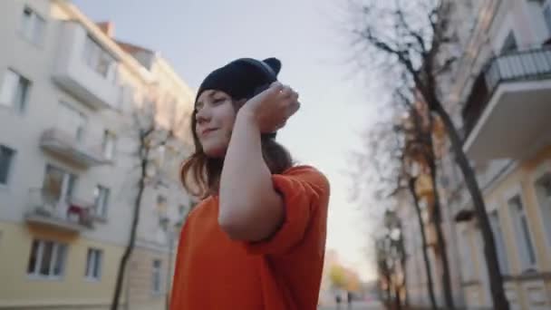 Linda chica joven caminando por la calle de la ciudad vieja y escuchar música en auriculares, estilo urbano, adolescente hipster con estilo en sombrero negro escuchar música y sonrisa, estilo naranja loco — Vídeos de Stock