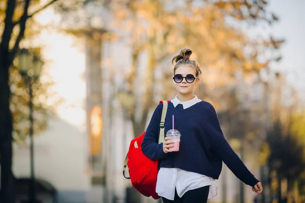 Adolescente muito hipster com saco vermelho bebe milkshake de uma xícara de plástico andando rua entre edifícios. Menina bonito em óculos de sol bebe uma bebida através de uma palha . — Fotografia de Stock