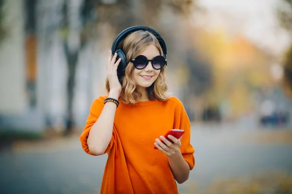 Aranyos fiatal lány sétál a régi város utca és a hallgató zene fejhallgató, városi stílusú, elegáns csípő tini hold mozgatható telefon és és őrült stílust mosoly, narancs — Stock Fotó