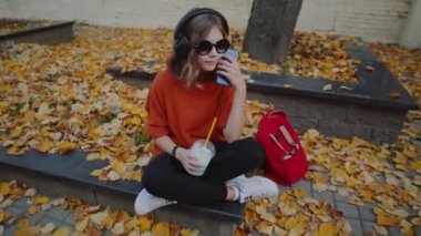 Güzel hipster teen dinlemek müzik içecekler milkshake binalar arasında street yürüyüş plastik bir kap dan kulaklık, kapatın. Güneş gözlüklü şirin kız bir saman içecek içecekler.