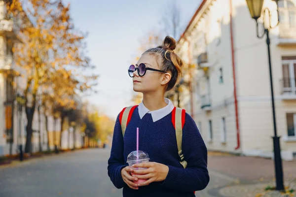 Adolescente muito hipster com saco vermelho bebe milkshake de uma xícara de plástico andando rua entre edifícios. Menina bonito em óculos de sol bebe uma bebida através de uma palha . — Fotografia de Stock