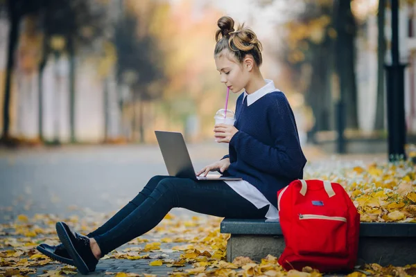 Sonbahar şehir sokak ve çalışma dizüstü bilgisayar kaldırımda oturan güzel hipster genç kız. Defter sıklıkta ve drinkking bir içecek bir saman kullanarak kız öğrenci. Güzel sonbahar hava.
