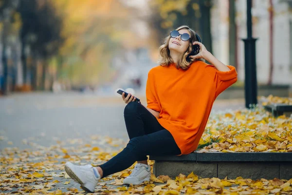 Милая молодая девушка слушает музыку в наушниках, в городском стиле, стильный хипстерский подросток сидит на тротуаре на городской улице и выбирает трек на мобильно перед зданиями одуванчивости, оранжевая улица — стоковое фото