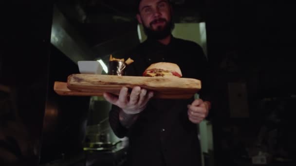 Sorrindo chef mestre em uniforme olhando para a câmera, segurando prato com hambúrguer servido . — Vídeo de Stock