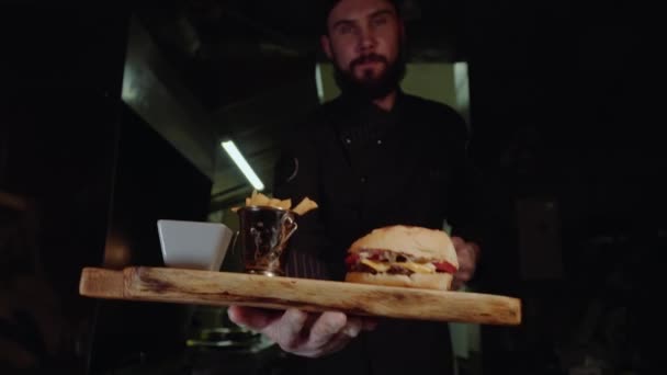 Chef maestro sonriente en uniforme mirando a la cámara, sosteniendo el plato con hamburguesa servida . — Vídeos de Stock