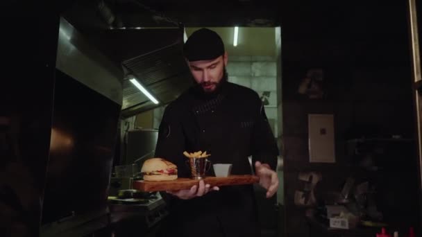 Sorrindo chef mestre em uniforme olhando para a câmera, segurando prato com hambúrguer servido . — Vídeo de Stock