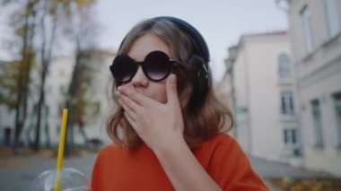 Güzel hipster teen dinlemek müzik içecekler milkshake binalar arasında street yürüyüş plastik bir kap dan kulaklık, kapatın. Güneş gözlüklü şirin kız bir saman içecek içecekler.
