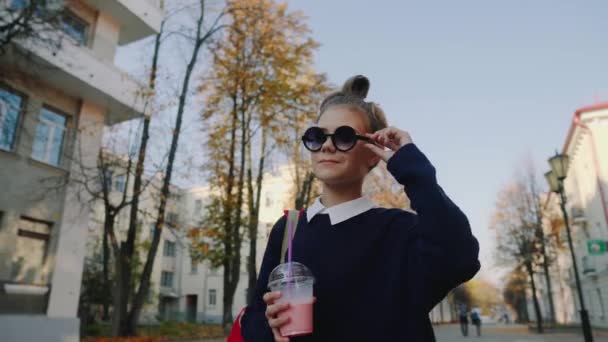 Bastante hipster adolescente con bolsa roja bebe batido de una taza de plástico caminando calle entre edificios. Linda chica en gafas de sol bebe una bebida a través de una paja . — Vídeo de stock