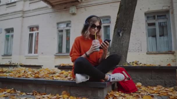 Jolie jeune fille écouter de la musique dans les écouteurs, style urbain, élégant hipster adolescent assis sur un trottoir sur la rue de la ville et de choisir la piste sur mobile infront de bâtiments oidsity, rue orange — Video