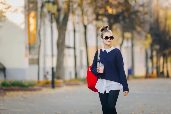 赤いバッグとかなり流行に敏感なティーンは、プラスチック カップの建物の間の通りを歩いてからミルクセーキを飲みます。サングラスでかわいい女の子ドリンク ストローで飲む. — ストック写真