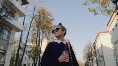 Güzel hipster teen kırmızı çanta ile bina arasında street yürüyüş plastik bir kap dan muzlu süt içiyor. Güneş gözlüklü şirin kız bir saman içecek içecekler.