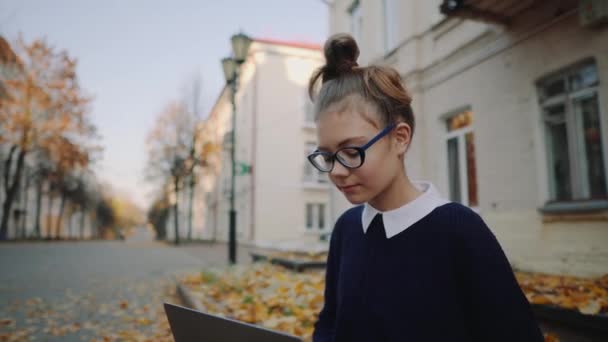 关闭漂亮的时髦的青少年女孩坐在人行道上的秋天城市街道和工作笔记本电脑。学生使用笔记本户外。美丽的秋天天气. — 图库视频影像