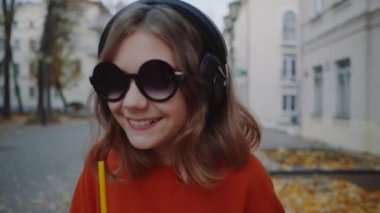 Güzel hipster teen dinlemek müzik içecekler milkshake gülümseyen ve binalar arasında street yürüyüş plastik bir kap dan kulaklık, kapatın. Güneş gözlüklü şirin kız bir saman içecek içecekler.