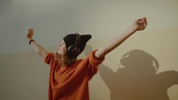 Schattig jong meisje luisteren muziek in hoofdtelefoons en dansen, stedelijke stijl, stijlvolle hipster tiener in zwarte hoed luisteren muziek en glimlach infront van gele muur, oranje gekke Streetstyle — Stockvideo