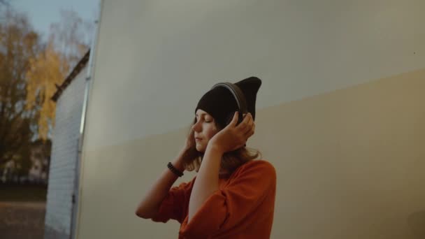 Niedliches junges Mädchen, das Musik im Kopfhörer hört und tanzt, urbaner Stil, stylischer Hipster-Teenager mit schwarzem Hut hört Musik und lächelt vor gelber Wand, orangefarbener verrückter Straßenstil — Stockvideo