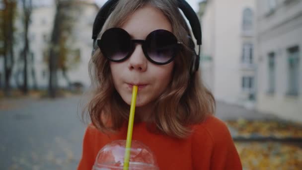 Close up Bastante hipster adolescente escuchar música en los auriculares, bebidas batido de una taza de plástico, sonriendo y caminando calle entre edificios. Linda chica en gafas de sol bebe una bebida a través de una paja . — Vídeo de stock