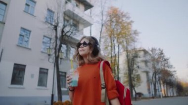 Güzel hipster teen içecekler milkshake plastik bir kap, gülümseyerek ve binalar arasında street yürüyüş üzerinden müzik kulaklık, dinle. Güneş gözlüklü şirin kız bir saman içecek içecekler.
