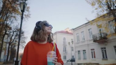 Güzel hipster teen içecekler milkshake plastik bir kap, gülümseyerek ve binalar arasında street yürüyüş üzerinden müzik kulaklık, dinle. Güneş gözlüklü şirin kız bir saman içecek içecekler.