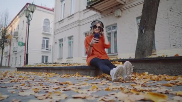 Bonito jovem ouvindo música em fones de ouvido, estilo urbano, elegante adolescente hipster sentado em uma calçada na rua da cidade e escolhendo faixa no celular na frente de edifícios antigos, rua laranja — Vídeo de Stock