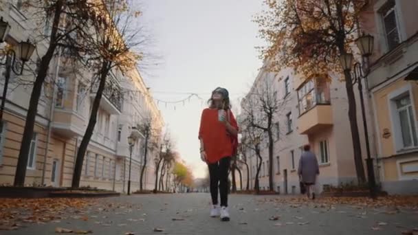 Bastante hipster adolescente escuchar música en los auriculares, bebidas batido de una taza de plástico, sonriendo y caminando calle entre edificios. Linda chica en gafas de sol bebe una bebida a través de una paja . — Vídeo de stock