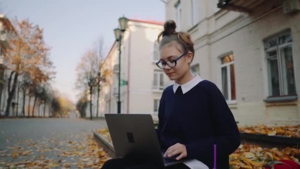 关闭漂亮的时髦的青少年女孩坐在人行道上的秋天城市街道和工作笔记本电脑。学生使用笔记本户外。美丽的秋天天气. — 图库视频影像