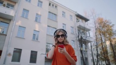 Sevimli Genç kız dinleme müzik kulaklık, kentsel tarzı, şık hipster teen mobilephone ve gülümseme oidsity binalar, turuncu çılgın sokak tarzı infront parça seçimi