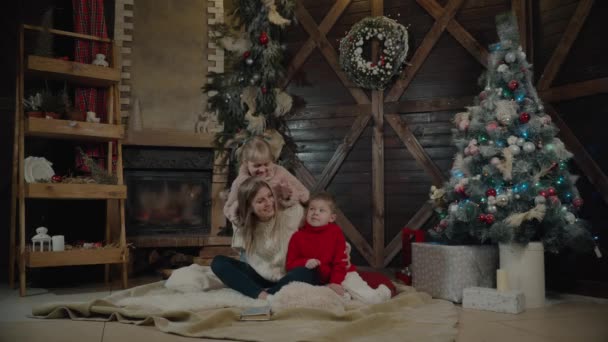 メリー クリスマスと新年あけましておめでとうございます。クリスマス インテリアに美しい家族です。かなり若い母親は彼女の娘と息子がクリスマス ツリーの近くに本を読んで. — ストック動画