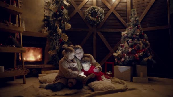 기쁜 성 탄과 새 해 복 많이 받으세요입니다. 크리스마스 인테리어에 아름 다운 가족입니다. 꽤 젊은 어머니 그녀의 딸과 아들 크리스마스 트리 근처 책을 읽고. — 비디오
