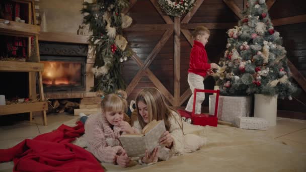 Feliz Natal e Feliz Ano Novo. Bela família no interior de Natal. Uma jovem mãe bonita a ler um livro para a filha. Filho decorar árvore de Natal . — Vídeo de Stock