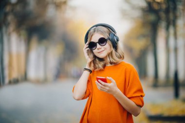 Sevimli Genç kız eski şehir sokak ve dinleme müzik kulaklık, kentsel tarzı, şık hipster teen tutun cep telefonu aşağı yürüyüş ve ve gülümseme, turuncu deli tarzı